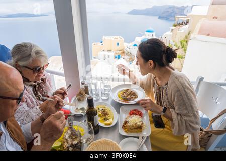 Tourists enjoy a mediterranean Greek lunch on a terrace restaurant overlooking a Greek island caldera, sharing local dishes, wine, and sunny seaside v Stock Photo
