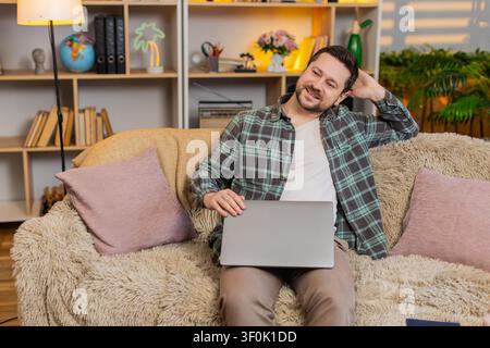 Young man on home sofa closes laptop after work and leans back with relieved smile after tasks. Adult guy on home sofa enjoys quiet rest and feels sat Stock Photo