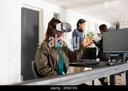 Black female developer with VR glasses, interacting with 3D coding interface. Woman analyzes explores virtual technology, demonstrating innovative software development at startup tech company. Stock Photo