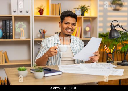 Indian man on home table reviews documents and sips coffee while checking paperwork and solving office tasks. Arabian guy on home table studies pages with calm focus during remote work and planning. Stock Photo