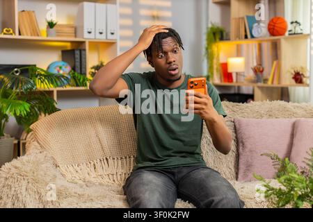 African American young man on home sofa looking at smartphone with sad face after bad news and online game loss. Black guy on home sofa feels upset while reading screen and reacting to online defeat. Stock Photo
