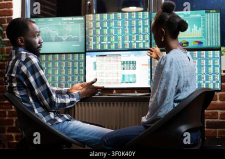 African American man and woman work late in an office, analyzing stock market trends and financial charts across multiple screens, linking stock trends and multi monitor. Stock Photo