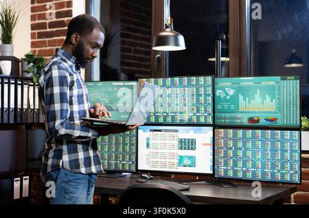 Financial analyst reviewing currency charts on laptop, black man standing and analyzing investment data on multiple screens in modern workstation. Stock Photo