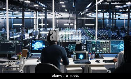 Software programmer enters a server room office and uses AI diagnostics to review infrastructure performance in a high tech facility. Stock Photo