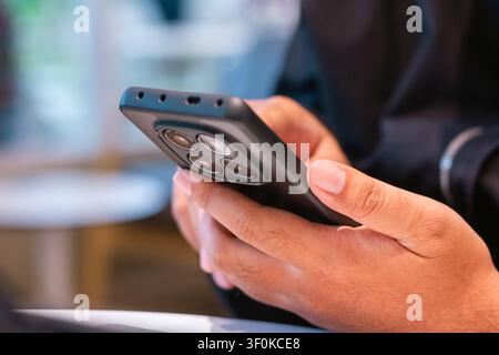 This close up image features a pair of hands holding a sleek mobile device with three distinct camera lenses, the background softly blurred to highlig Stock Photo