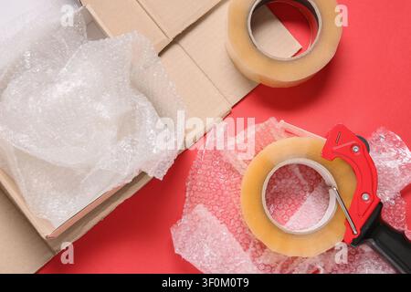 Shipping supplies with bubble wrap, cardboard box, and packing tape on a red background, top view Stock Photo