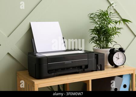 Black inkjet printer with blank paper on a wooden shelf next to a potted plant and alarm clock, the background features a sage green wall with a subtl Stock Photo