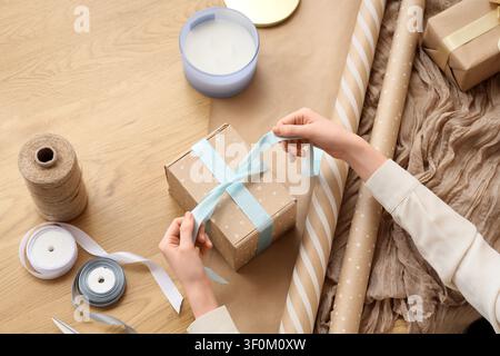 Person tying a light blue ribbon around a polka dot kraft paper gift box on a wooden table with wrapping supplies, the wooden table is filled with var Stock Photo