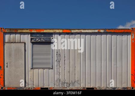 Weathered Portable Construction Site Office Container with Closed Roller Shutter Against Blue Sky Stock Photo