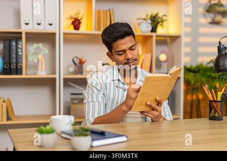 Indian man on home table reads book makes notes and pauses to think during focused study indoors at home. Arabian guy on home table reviews pages and writes ideas for learning planning and home work. Stock Photo