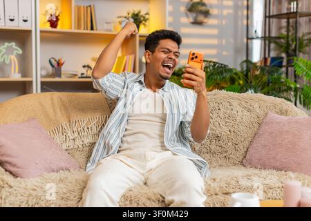 Indian man on home sofa checks smartphone and celebrates personal win with raised hand and excited smile in living room. Arabian guy on home sofa reads good news on phone and enjoys victory at home. Stock Photo