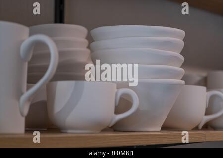 Stacked white ceramic cups and bowls displayed on a wooden shelf in a modern kitchen or cafe interior. The clean minimalist composition represents kit Stock Photo