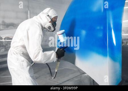 Worker in protective mask and jumpsuit overall spraying base coat of paint on car body elements in painted chamber using professional airbrush. Stock Photo