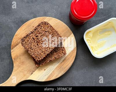 Sliced dark seed bread on a wooden cutting board with butter and a jar of red jam placed on a gray surface Stock Photo