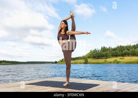 Woman doing standing vertical splits yoga pose Urdhva Prasarita Eka Padasana on a pier by the lake at sunny summer morning Stock Photo