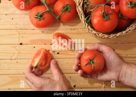 Close-up of fresh ripe tomatoes on a wooden table, with hands holding whole and sliced tomatoes. Rustic kitchen scene with natural light and harvest c Stock Photo