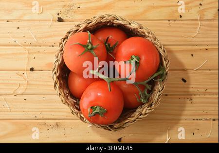Top view of a wicker basket filled with fresh red tomatoes on a rustic wooden table. Bright natural light emphasizes organic produce and harvest fresh Stock Photo