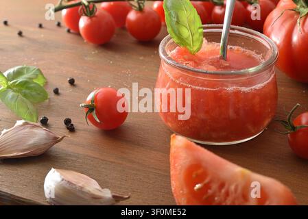 Close-up of homemade fresh tomato spread in a glass jar with basil, surrounded by cherry tomatoes, garlic, and rustic wooden table in warm natural lig Stock Photo
