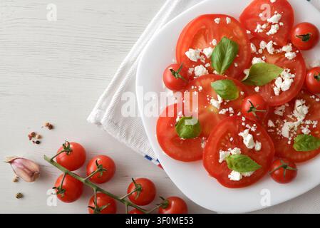 Top view of a fresh tomato salad with basil leaves and crumbled cheese on a white plate, surrounded by cherry tomatoes, garlic, and spices on a light Stock Photo