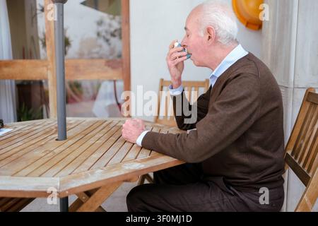 Senior man sitting outdoors using an asthma inhaler for breathing Stock Photo
