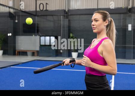 Woman holding black paddle and watching yellow ball on blue court wearing pink tank, copy space. Padel, racket, practice, female, facility, modern, focused Stock Photo