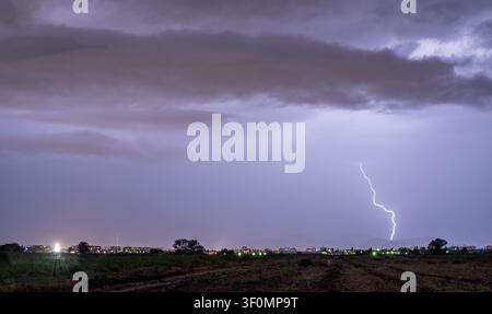 A powerful cloud-to-ground lightning bolt strikes near the outskirts of a mid-sized city, captured from an open agricultural flatland at dusk. Stock Photo