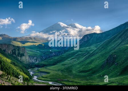 Snow-capped Mount Elbrus, the highest peak in Europe at 5,642 meters, rises above the lush green Baksan Valley in Kabardino-Balkaria, Russia. Stock Photo