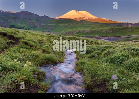 A glaciated twin-summit Mount Elbrus, Europe's highest peak at 5,642 meters, rises above lush summer alpine meadows in Kabardino-Balkaria, Russia. Stock Photo