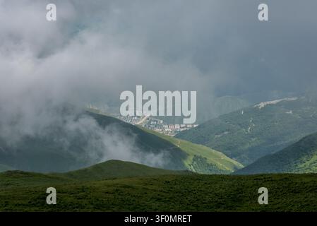 A sweeping aerial-perspective view across lush green alpine meadows toward a compact mountain town nestled deep in a Caucasus valley. Stock Photo