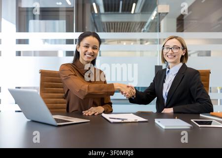 Two smiling multi-ethnic businesswomen shaking hands after a successful meeting, collaborating on a new project in a modern office, symbolizing agreement and closing a deal Stock Photo