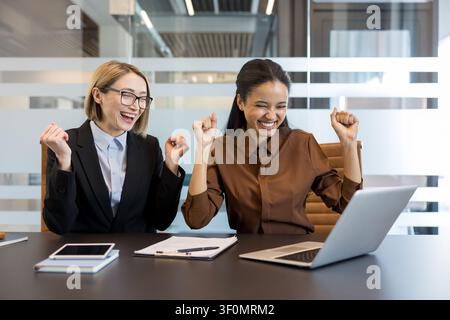 Two diverse businesswomen sitting at a desk, looking at a laptop screen and cheering with excitement, actively celebrating a shared success or milestone in a corporate office setting Stock Photo