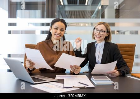 Two smiling businesswomen sitting at a desk and holding documents, celebrating a successful project completion in a contemporary corporate office environment Stock Photo