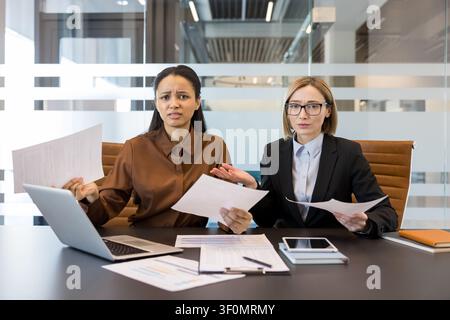 Two stressed businesswomen sit at a modern office desk, holding papers and looking frustrated, struggling with complex documents and facing work challenges Stock Photo