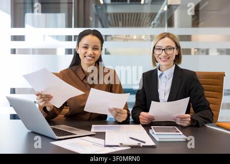 Diverse businesswomen smiling at camera while working together on documents and a laptop in a contemporary office setting, demonstrating teamwork and professional cooperation Stock Photo
