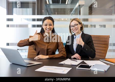 Two cheerful businesswomen smiling and giving thumbs up, celebrating success and agreement during a productive meeting in a modern office environment Stock Photo