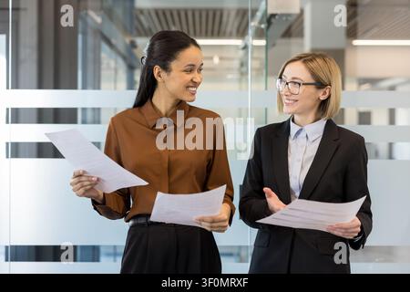Two diverse businesswomen smiling and discussing paperwork, collaborating on a project in a modern office, representing teamwork, partnership, and professional communication Stock Photo