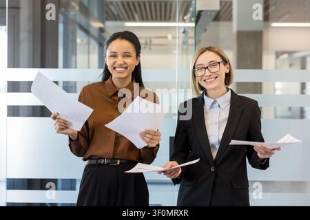 Two diverse businesswomen smiling confidently, holding important documents and discussing successful project results in a modern corporate office environment Stock Photo