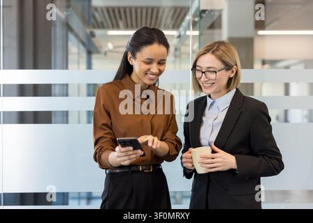 Two smiling multi-ethnic businesswomen are sharing content on a smartphone and drinking coffee during a casual conversation in a contemporary office setting, representing teamwork and cooperation Stock Photo