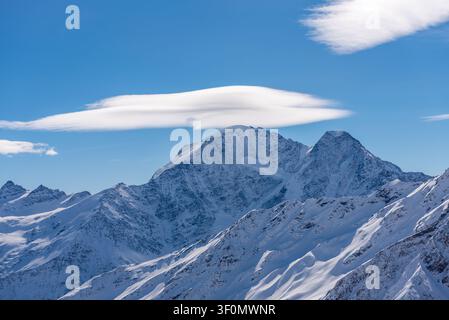 A towering snow-covered peak in the Greater Caucasus mountain range rises prominently above a layered series of ridges and glaciated slopes. Stock Photo