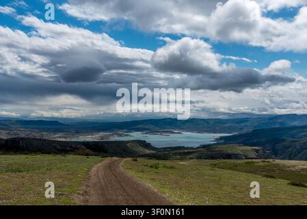 A winding unpaved dirt road descends across a sweeping green highland plateau toward a vivid turquoise mountain reservoir framed by layered cliff esca Stock Photo