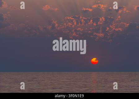 A vivid red solar disk hangs just above the horizon over a calm, glassy sea. Stock Photo