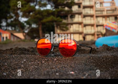 Round aviator-style sunglasses with red mirrored lenses rest on dark coarse sand of a Black Sea resort beach. Stock Photo