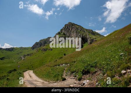 A winding unpaved dirt road curves through a lush alpine valley beneath a dramatic rocky limestone peak in the Greater Caucasus mountain range. Stock Photo