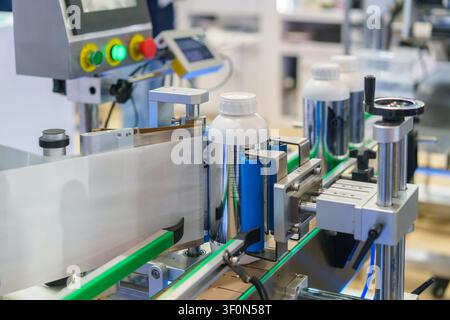 Close-up of an automatic labeling machine applying white paper labels to plastic bottles moving along a conveyor belt in a modern manufacturing plant. Stock Photo
