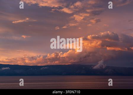 A wide panoramic view across the calm surface of Lake Baikal (Ozero Baykal) toward the darkened mountain ridge of the opposite shore. Stock Photo