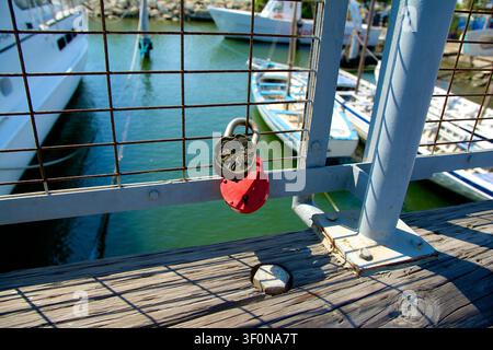 A weathered red padlock hangs on a rusty metal chain fence overlooking a picturesque marina filled with colorful fishing and leisure boats. Stock Photo