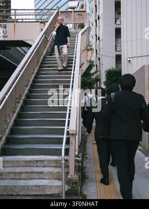 Man descending a pedestrian footbridge staircase while businessmen in suits walk along a sidewalk in Shibuya, Tokyo, Japan Stock Photo