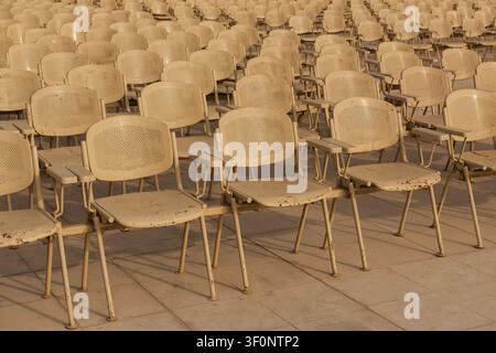 Vast rows of empty weathered cream metal chairs filling outdoor stone plaza bathed in warm amber sunlight ideal for event capacity solitude and public Stock Photo