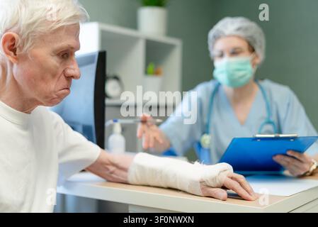 Senior patient with arm cast writing notes during medical visit. Concerned older gentleman documenting treatment details while doctor observes. Involved elderly man in own healthcare recovery Stock Photo