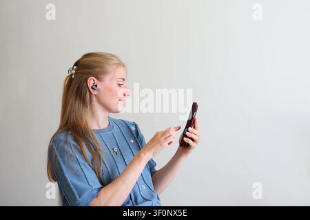 Young woman using wireless earbuds while operating a smartphone, enjoying hands-free communication and mobile technology. Stock Photo